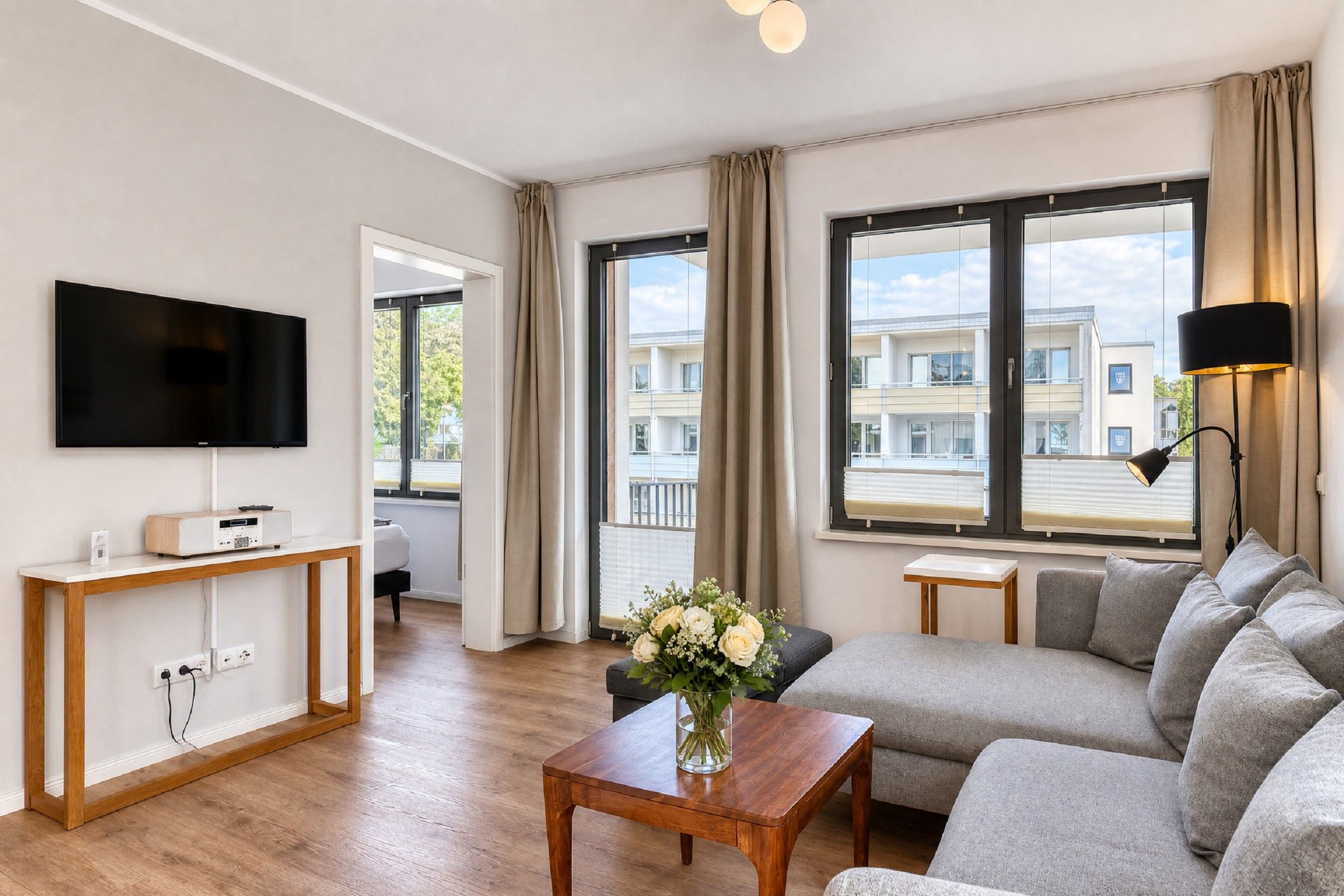 A modern living room with a flat-screen TV, a wooden coffee table with a vase of flowers, a grey sofa, and a view of a building through large windows.