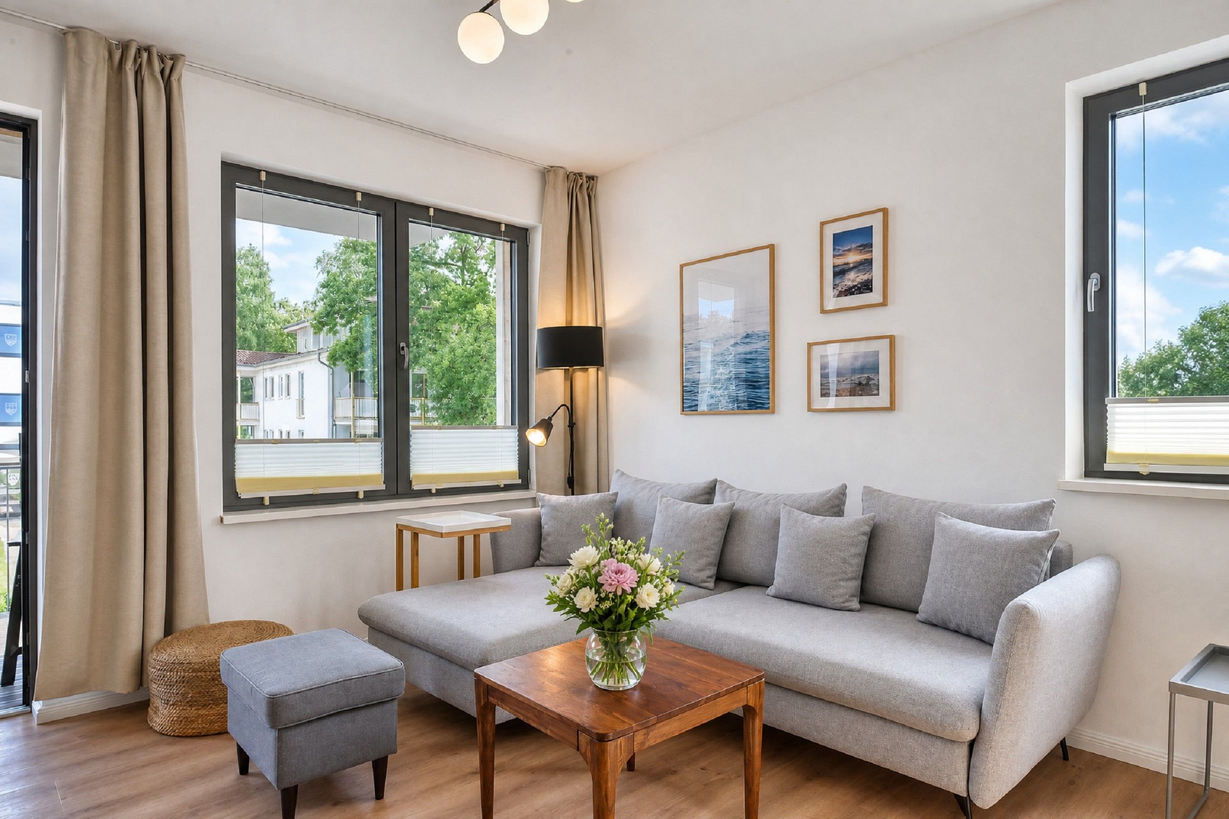 Una sala de estar moderna con un sofá gris, una mesa de centro de madera y un pequeño taburete. Hay dos ventanas con cortinas, una lámpara de pared y tres cuadros enmarcados.