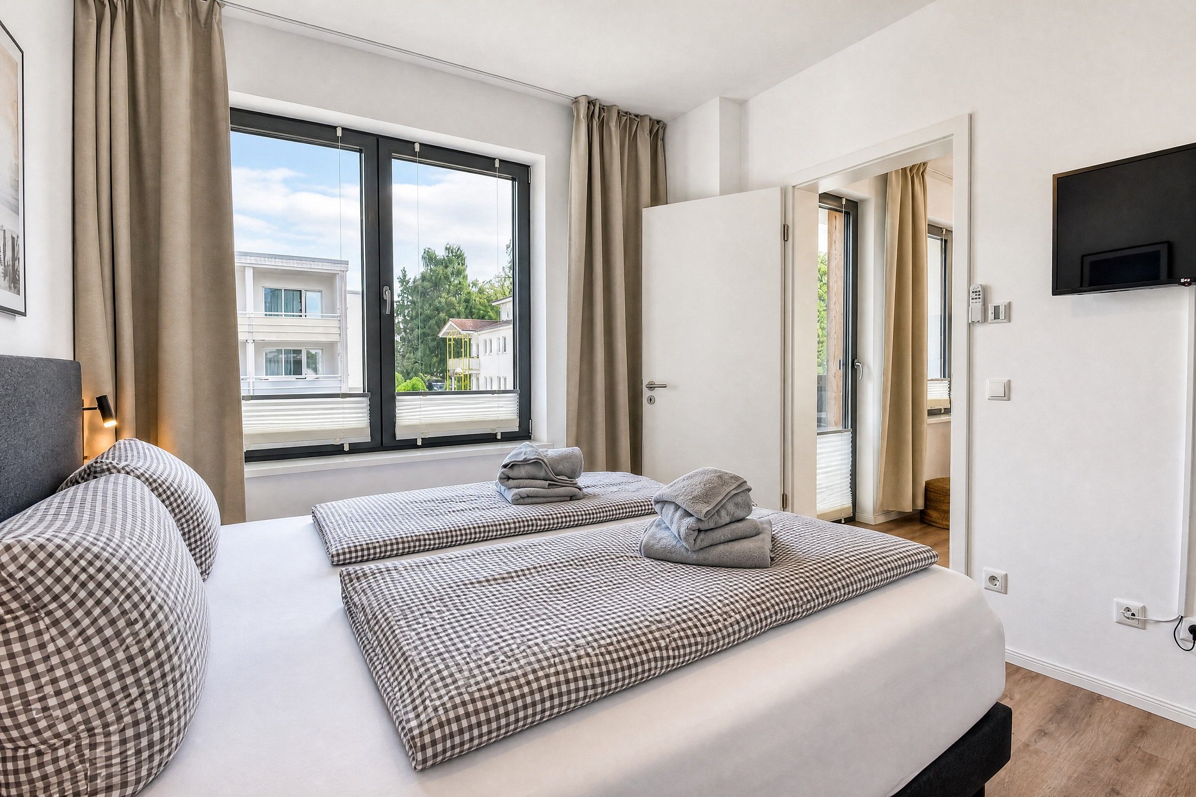 A hotel room with a large window, a bed with checkered bedding, a flat-screen TV mounted on the wall, and a door leading to a balcony.