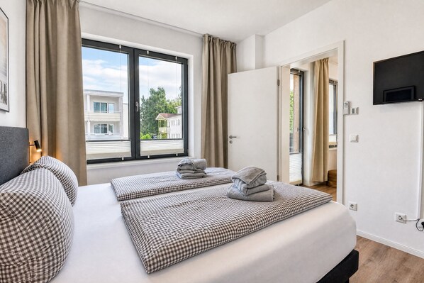A hotel room with a large window, a bed with checkered bedding, a flat-screen TV mounted on the wall, and a door leading to a balcony.