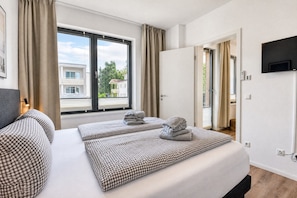 Habitación de hotel con un ventanal grande, cama con ropa de cama a cuadros, televisor de pantalla plana montado en la pared y puerta que da a un balcón.