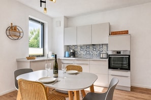 A modern kitchen with white cabinets, a built-in oven, and a dining area with a round table and wicker chairs.
