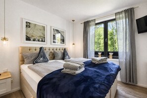 Un dormitorio con cama, dos cuadros enmarcados, una ventana con cortinas, una mesita de noche y un televisor de pantalla plana en la pared.