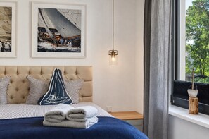 A bedroom with a tufted headboard, a bed with checkered pillows, a sailboat-themed pillow, and a stack of folded towels. There are two framed pictures on the wall, a hanging light fixture, and a window with curtains.