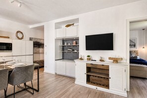 A modern living room with a flat-screen TV mounted on the wall, a dining area with a table set for two, and a cozy seating area with a sofa.