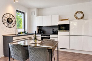 A modern kitchen with white cabinets, a dining table with chairs, a clock, and a window with a view of trees.