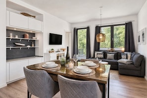 Eine moderne Küche mit eingebauten Regalen, einem Esstisch für vier Personen und einem Wohnbereich mit Sofa und großen Fenstern.