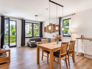 A modern living space with a dining area, sofa, and large windows offering a view of greenery.