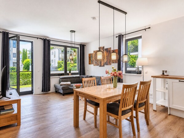 A modern living space with a dining area, sofa, and large windows offering a view of greenery.