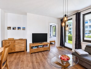 A modern living room with a TV, wooden furniture, a glass table with apples, and a view of a building outside.