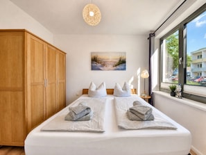 A bedroom with a bed, a wooden wardrobe, a painting, a window with a view, and a lamp.