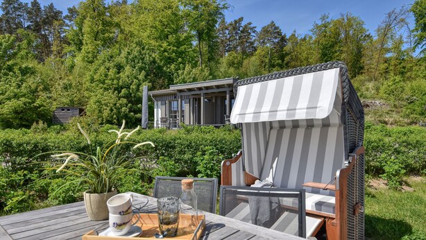A wooden deck with outdoor furniture, a dining table with a tray, and a house with a balcony in a forested area. - Newly built, modern, exclusive - that describes the Klein Stresow vacation village (Klein Stresow)