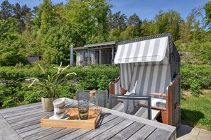 Una terraza de madera con mobiliario exterior, una mesa de comedor con una bandeja, y una casa con balcón en un área boscosa.
