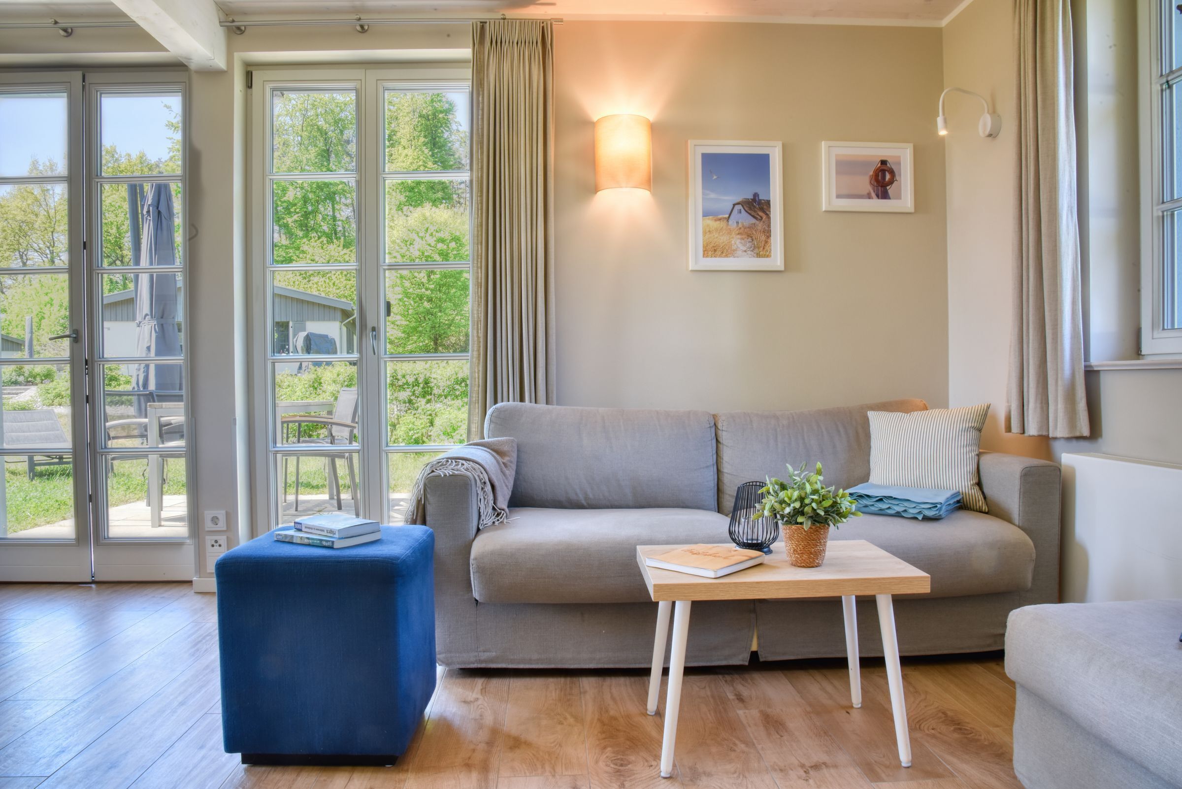 Una sala de estar con un sofá, una mesa de centro y un reposapiés azul. Hay dos ventanas con cortinas, una lámpara de pared y dos cuadros enmarcados.