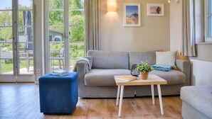 A living room with a sofa, a coffee table, and a blue ottoman. There are two windows with curtains, a wall-mounted light, and two framed pictures. - Newly built, modern, exclusive - that describes the Klein Stresow vacation village (Klein Stresow)