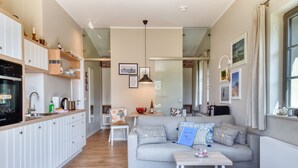 Una cocina moderna con armarios blancos, encimera de madera y horno empotrado. Hay un comedor con una mesa y sillas, y una sala de estar con un sofá gris y una mesita de centro.