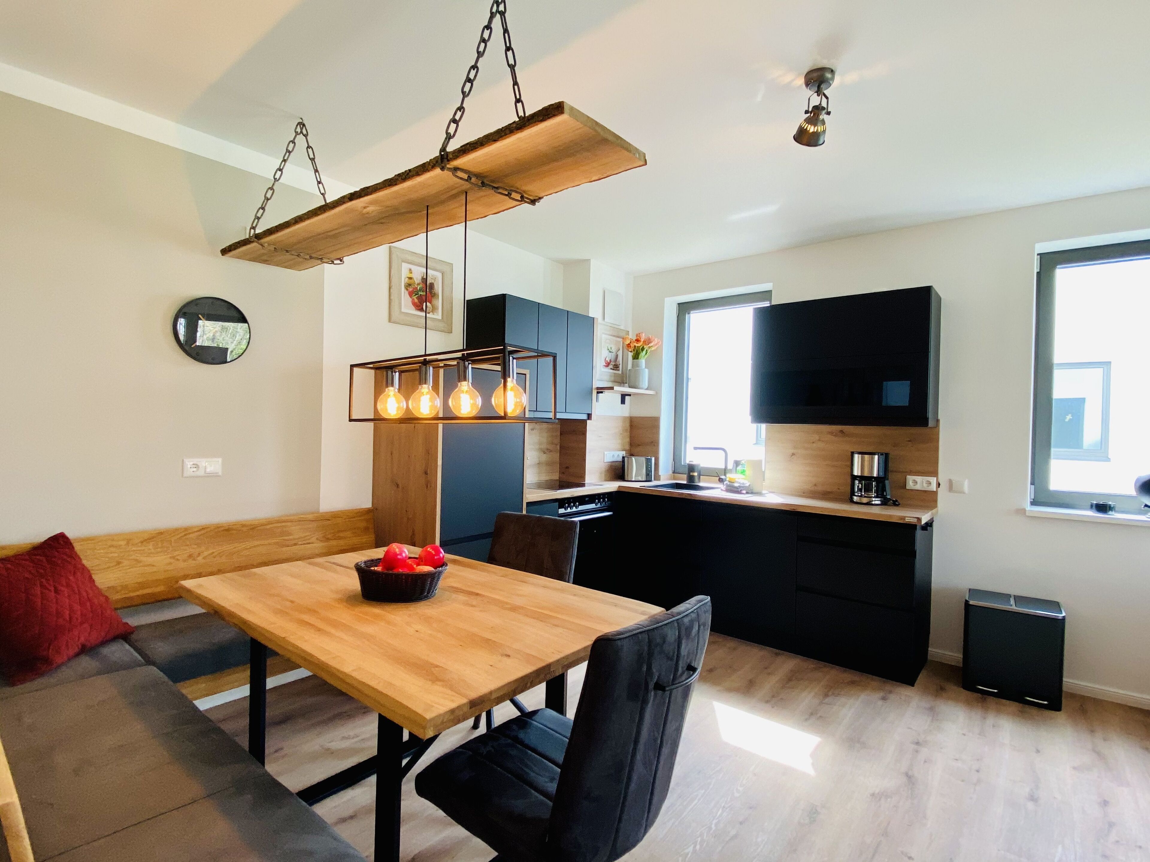 Una cocina moderna con encimera de madera, armarios negros y horno empotrado. Incluye un comedor con mesa de madera y sillas negras.