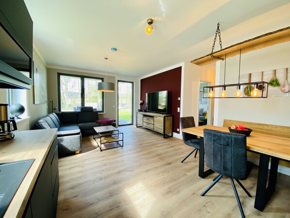 Een moderne woonkamer met een bank, salontafel en een eethoek met stoelen. Er is een televisie, een keuken met kasten en een groot raam met uitzicht op de omgeving.