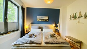 A neatly made bed with checkered bedding, a bedside table with a lamp, a painting on the wall, and a bench by the window.