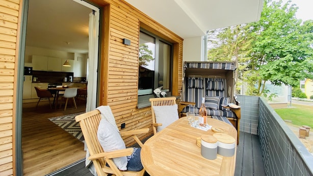 A wooden deck with a round table and chairs, a sliding glass door leading to a kitchen, and a view of a tree and a fence.