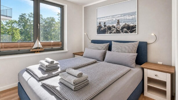 Un dormitorio con un ventanal grande, una cama con ropa de cama a cuadros, una mesita de noche y un cuadro colgado en la pared.