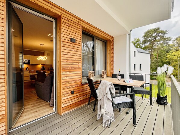 Una terraza de madera con una mesa y sillas de comedor, un área de descanso acogedora con cojines, y vistas a un jardín y un edificio blanco al fondo.