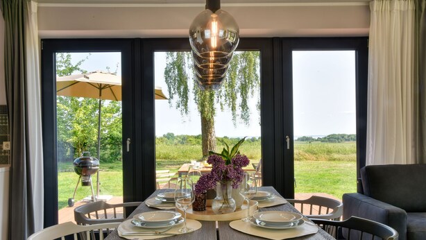 A dining area with a table set for six, a hanging light fixture, and a view of a grassy landscape through large windows. - Welcome to the Brick Quarter South Putbus on the island of Rügen (Putbus)