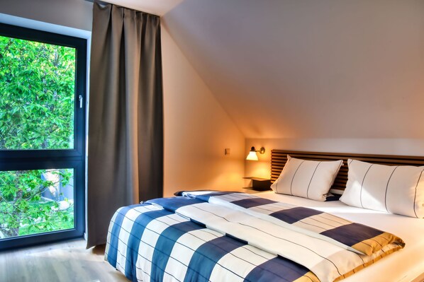 A hotel room with a large window, a bed with a striped bedspread, a nightstand with a lamp, and a view of greenery outside. - House half brick quarter west with sauna, fireplace and wonderful water view! (Putbus)