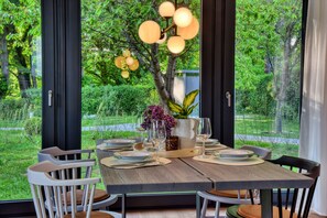 A dining table set for four with chairs, a vase with flowers, and a view of a lush garden through large windows. - House half brick quarter west with sauna, fireplace and wonderful water view! (Putbus)