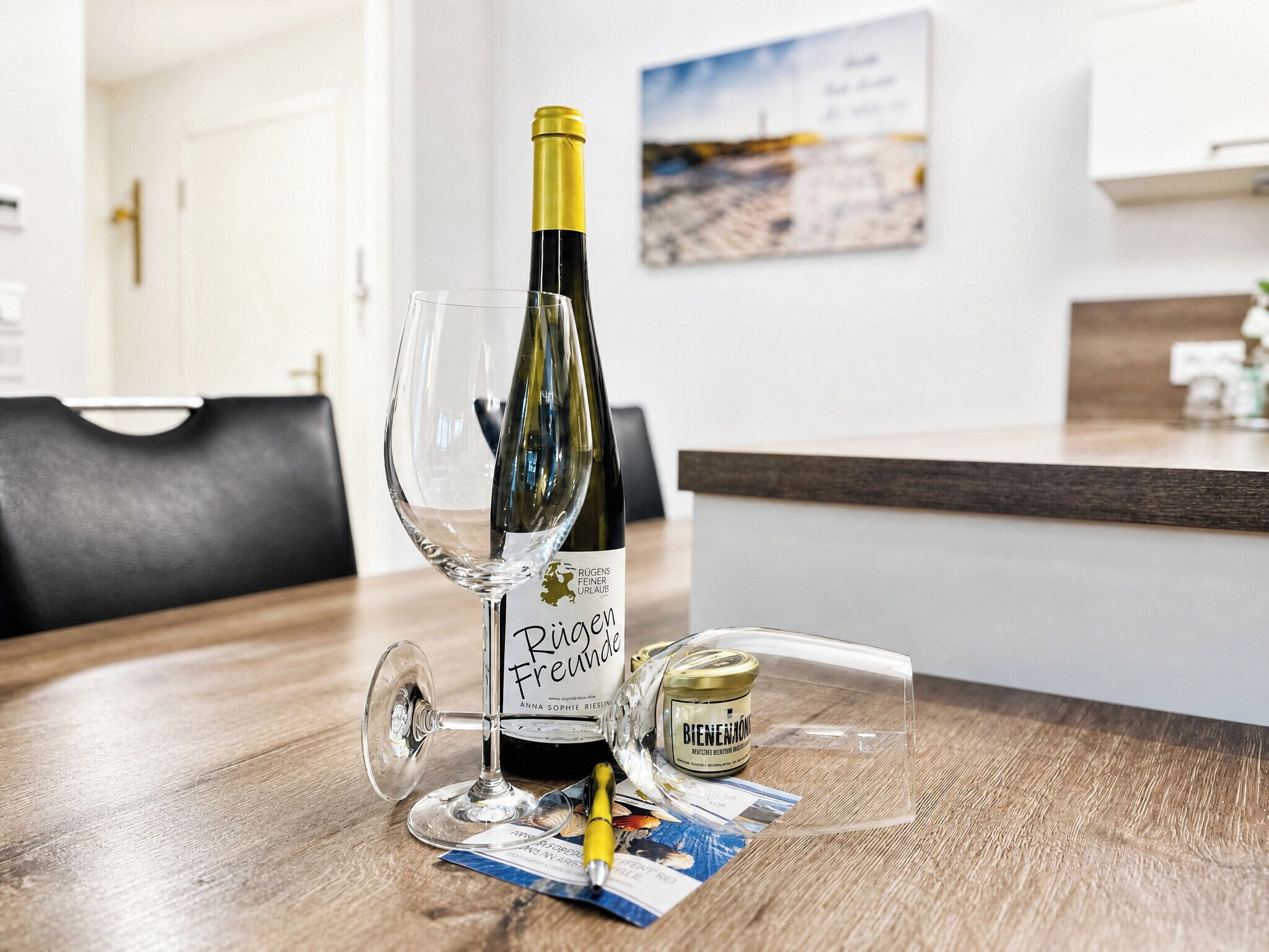 Une table en bois sur laquelle se trouvent une bouteille de vin, un verre à vin et un petit récipient, dans une pièce aux murs blancs ornée d’un tableau.