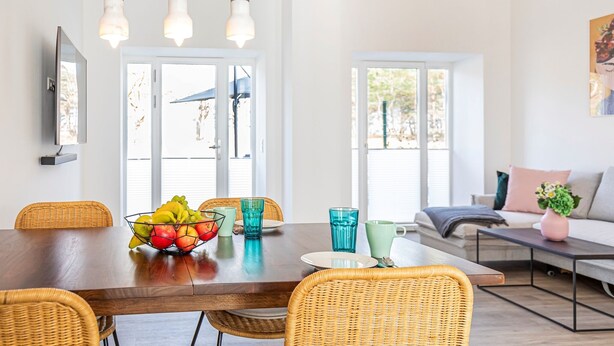 A dining area with a wooden table, wicker chairs, and a fruit bowl. There are large windows and a sofa in the background. - Sunshine by the sea: a modern oasis for unforgettable moments in Prora, Rügen (Prora)