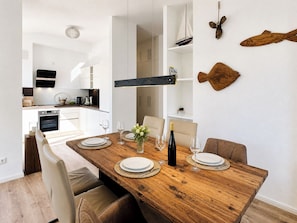 Cozinha moderna com armários brancos, sala de jantar com mesa de madeira para quatro pessoas e sala de estar com sofá e televisão.