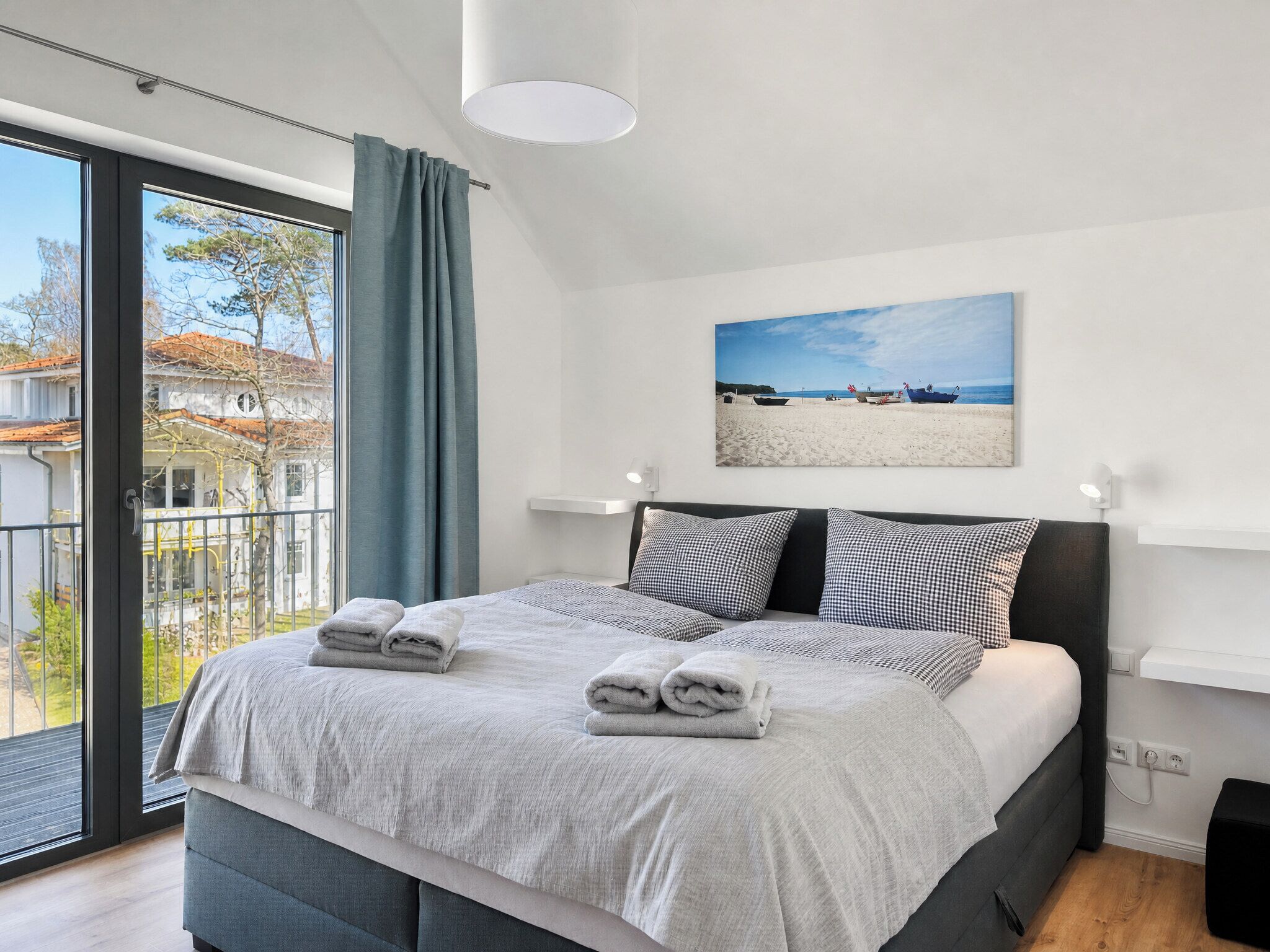 A modern bedroom with a large bed, a view of the outside, and a beach scene painting on the wall.
