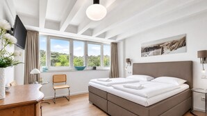 Un dormitorio con cama, escritorio, silla, ventana con cortinas, televisor montado en la pared y un cuadro.