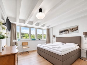 A bedroom with a bed, a desk, a chair, a window with curtains, a wall-mounted TV, and a painting.