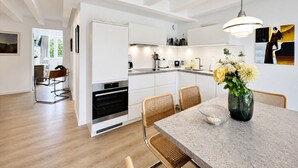 Una cocina moderna con armarios blancos, horno empotrado y un comedor con mesa de mármol y sillas de mimbre.