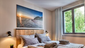Et soveværelse med et stort vindue, en seng med ternet sengetøj, et natbord med en lampe og et maleri af en solnedgang over vand.