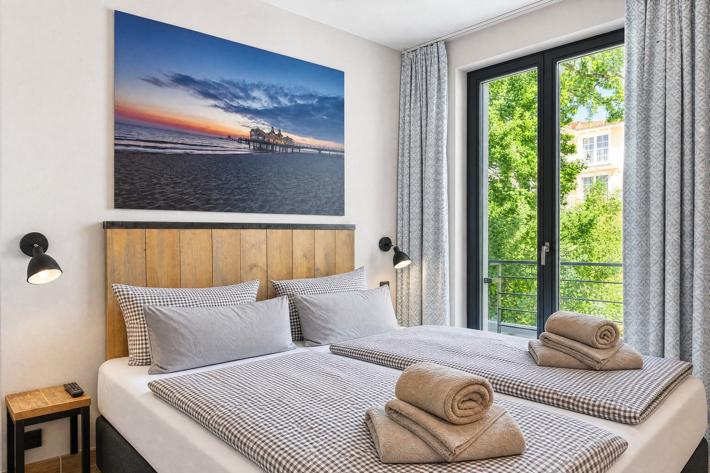A hotel room with two beds, a large window with curtains, a painting of a sunset, and a view of greenery outside.