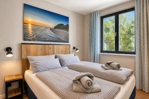 Una cama doble con ropa de cama y toallas a cuadros, un cabecero de madera, una mesita de noche, una lámpara de pared y un amplio ventanal con cortinas.
