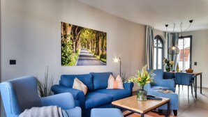 Un salon moderne avec un canapé bleu, une table basse en bois et un tableau représentant une route bordée d’arbres accroché au mur.