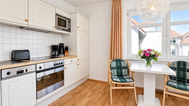 Una cocina moderna con armarios blancos, microondas integrado, estufa y tostadora. Cuenta con un comedor con mesa blanca, dos sillas y un florero con flores.