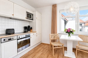 Una cocina moderna con armarios blancos, horno empotrado, microondas y cafetera. Hay una zona de comedor con una mesa blanca y dos sillas de mimbre. Se ve una ventana con vistas a edificios.