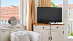 Una habitación con televisión sobre un soporte de madera, un armario blanco con cajones y una ventana con cortinas.