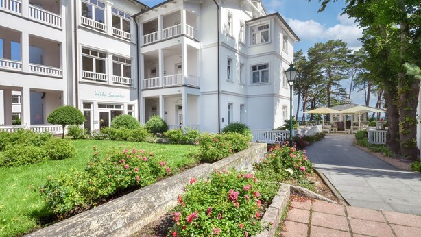 Un hotel de varios pisos con balcones, un jardín bien cuidado con flores rosadas y un camino pavimentado que conduce a la entrada.