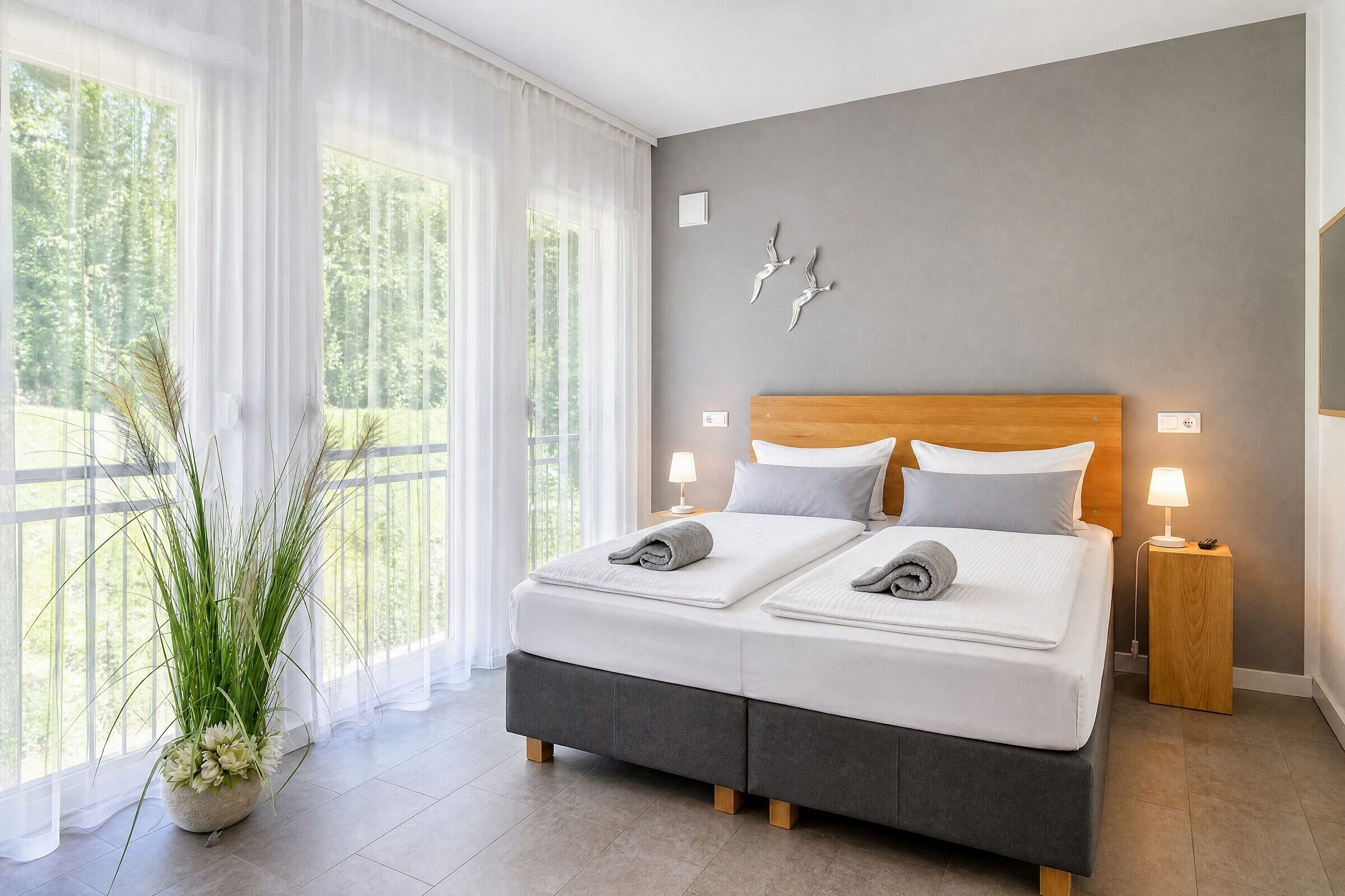 Un dormitorio moderno con una cama grande, dos mesitas de noche, una planta en maceta y vistas a zonas verdes por la ventana.