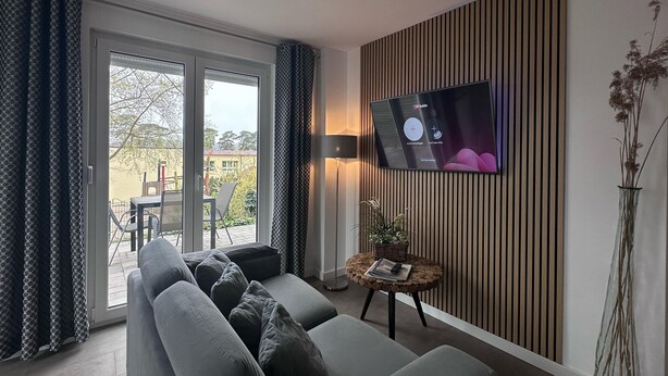 A modern living room with a grey sofa, a flat-screen TV mounted on the wall, a small round table with a lamp, and a view of the outdoors through a sliding glass door.