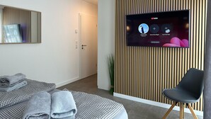 A modern hotel room with a flat-screen TV mounted on a wooden panel wall, a bed with a checkered bedspread, a black chair, and a mirror on the wall.