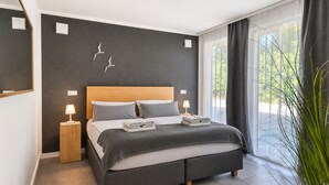 A modern bedroom with a large bed, bedside lamps, a wooden headboard, and a view of greenery through sheer curtains.