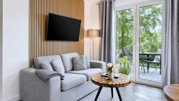 A modern living room with a grey sofa, a flat-screen TV mounted on a wooden panel wall, a round wooden coffee table with a vase of flowers, and a sliding glass door leading to an outdoor patio.
