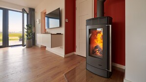 A modern living room with a wood-burning stove, a large flat-screen TV, and a sliding glass door leading to a balcony. - Welcome to Backsteinquartier B Putbus on the island of Rügen (Putbus)